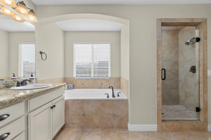 Built-in bathtub with tiled surround, separate glass-enclosed shower with tiled walls, dual-sink vanity with stone countertop, and wood-finish cabinetry