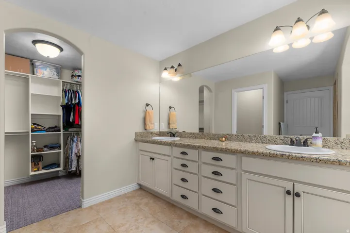 Dual vanity with granite-style countertop, integrated sinks, tile flooring, and arched entryway to a walk-in closet with built-in shelving