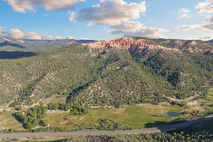 Expansive natural landscape with varied topography, featuring dense evergreen forests, red rock formations, a tranquil pond, and a paved roadway
