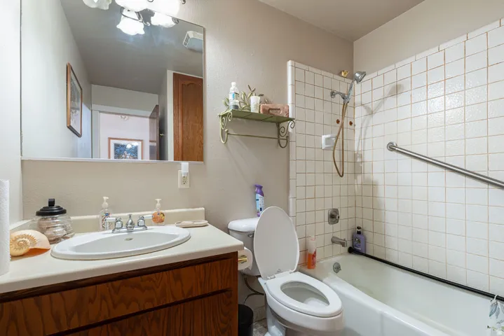 White tile shower with grab bar, white bathtub, vanity with wood-finish cabinetry, and an integrated sink with a chrome faucet