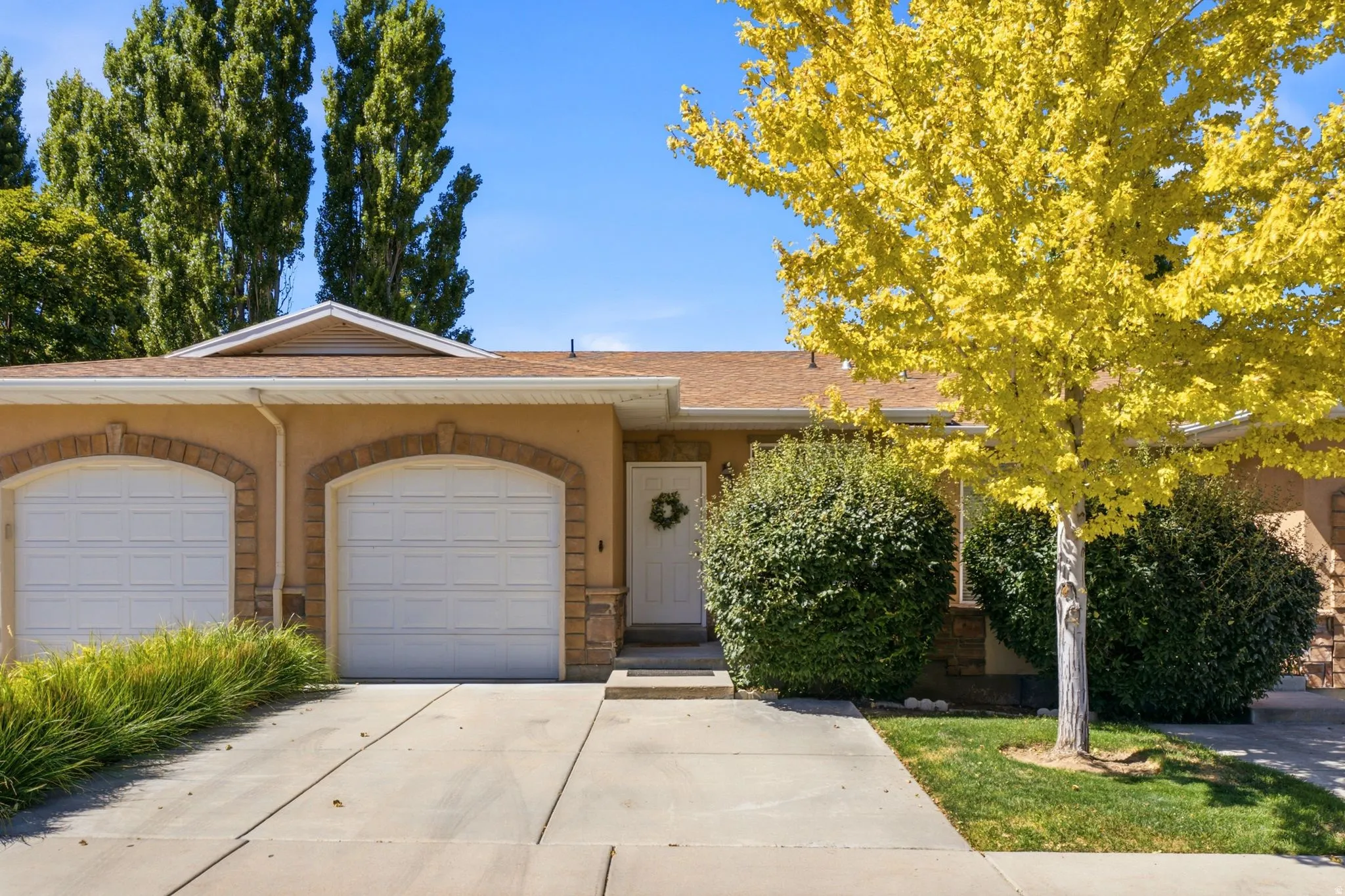 Tan stucco exterior with stone accents, featuring two arched garage doors, a white entry door, and mature landscaping