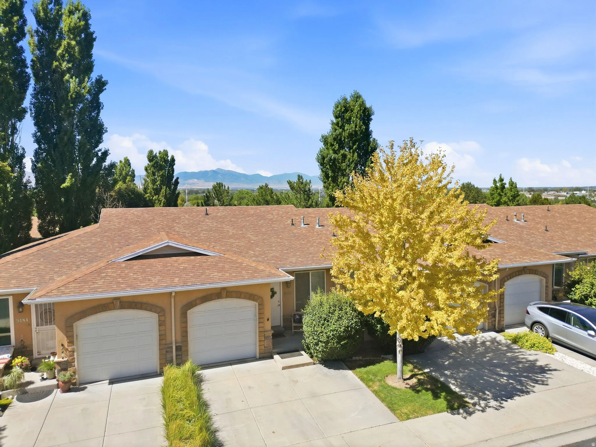 Duplex property featuring a brown shingle roof, arched garage door surrounds, a paved driveway, and mature landscaping
