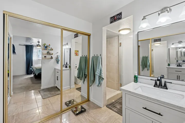 Bathroom with tile flooring, dual vanities featuring white cabinetry and black hardware, a vanity with a white countertop, a black-framed mirror, and a tiled shower stall