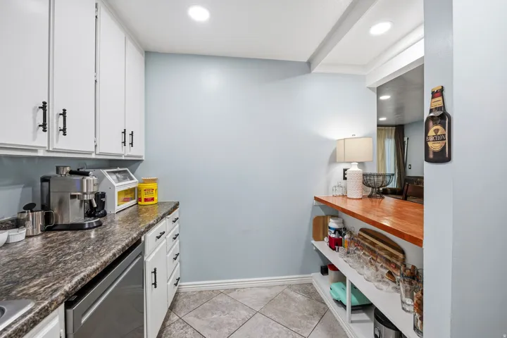 White cabinetry with matte black hardware, dark variegated countertops, stainless steel dishwasher, tile flooring, and a wood-finish breakfast bar with integrated shelving