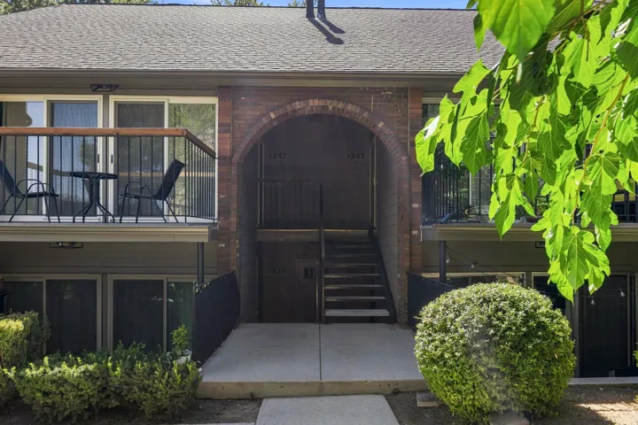 Brick archway entrance with a concrete walkway, exterior staircase, upper-level balcony, and mature landscaping