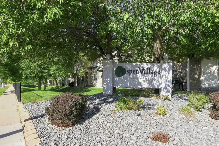 Aspen Village monument sign featuring mature trees, landscaped rock beds, and a paved sidewalk