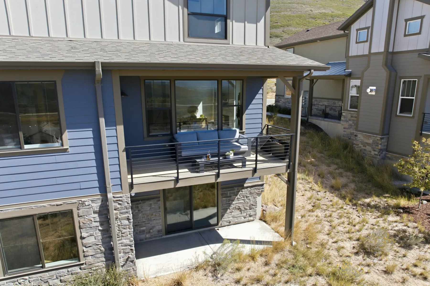 Exterior with blue horizontal siding, stone veneer accents, an upper-level deck with horizontal railing, and a ground-level concrete patio