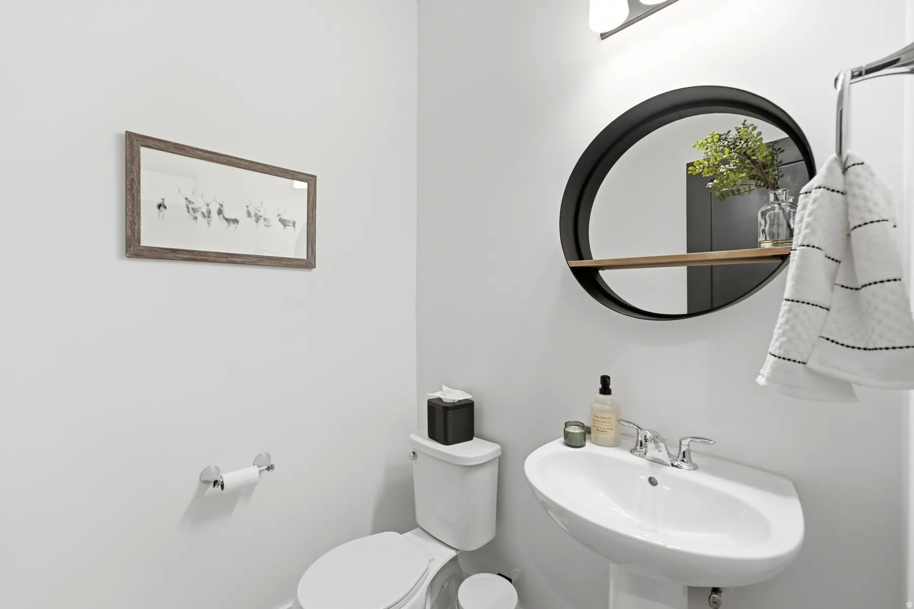 Pedestal sink with chrome faucet, wall-mounted circular mirror with integrated wood-finish shelf, and polished chrome towel bar