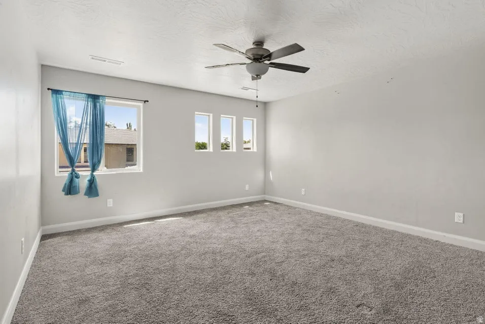 Spacious room featuring neutral carpeting, light-toned walls, multiple windows providing natural illumination, and a ceiling fan
