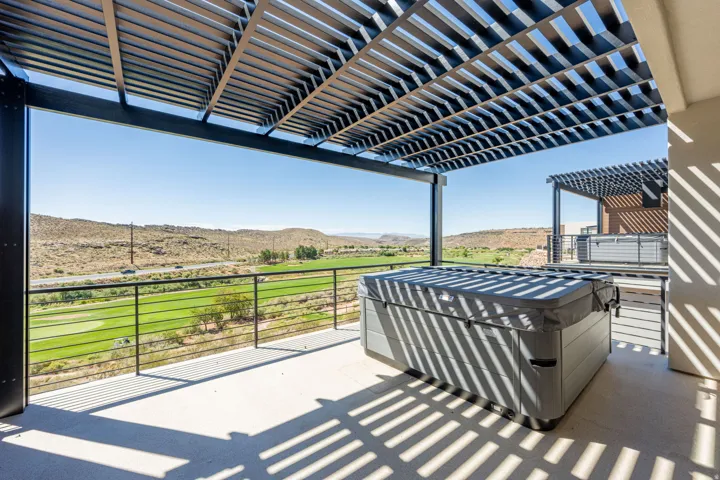 Covered outdoor patio with a hot tub, slat pergola, and metal railing overlooking a golf course and distant hills