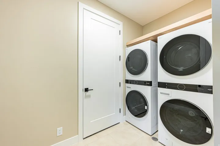 Hard-surface flooring, solid core door with matte black hardware, dual stackable laundry machines, and a wood-finish shelf