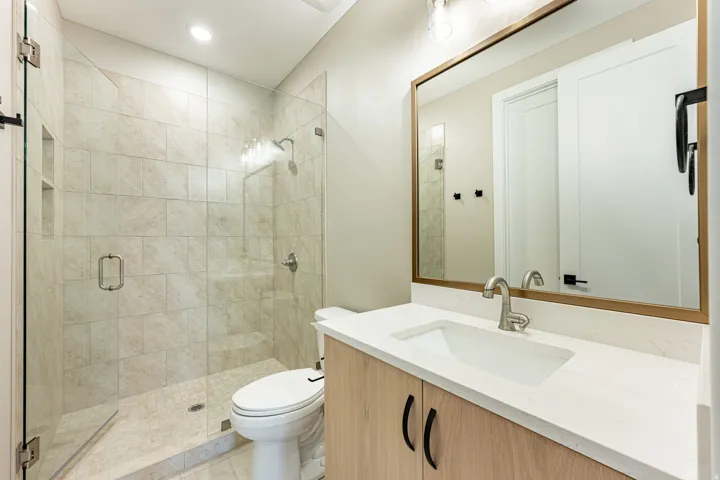 Frameless glass shower enclosure with tiled walls, wood-finish vanity with countertop, rectangular undermount sink, and a large framed mirror