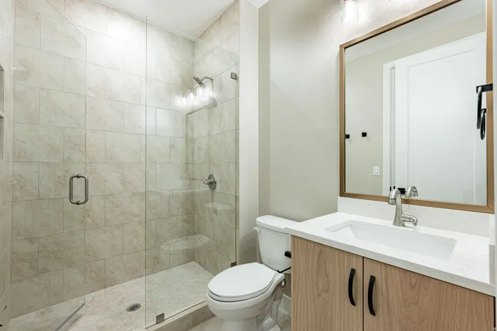 Glass-enclosed shower with tiled walls, wood-finish vanity with an integrated sink, quartz countertop, and a framed mirror with an overhead light fixture