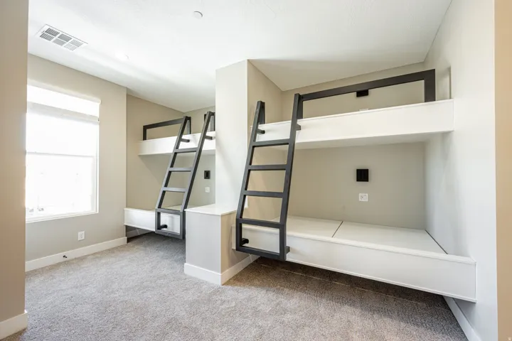 Built-in bunk beds with black metal ladders, neutral carpeting, and a window with a roller shade