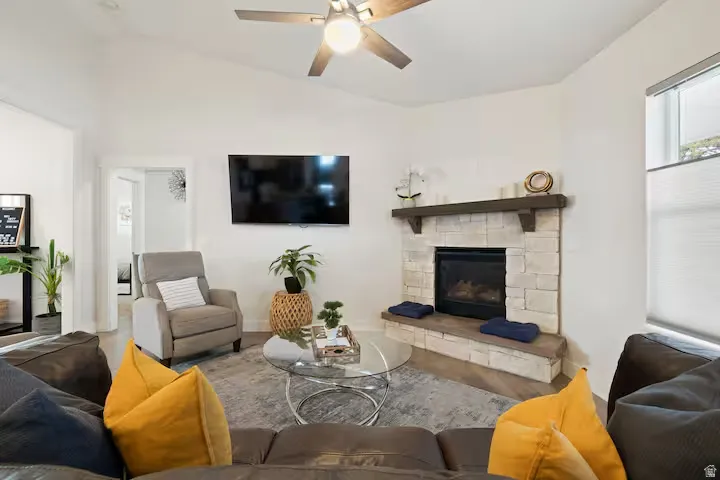 Vaulted ceiling with a ceiling fan, stone-clad fireplace with a wood mantle, and wood-finish flooring