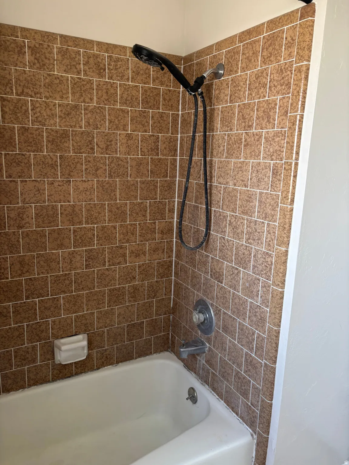 Bathtub with brown tiled surround, featuring a handheld showerhead and wall-mounted faucet