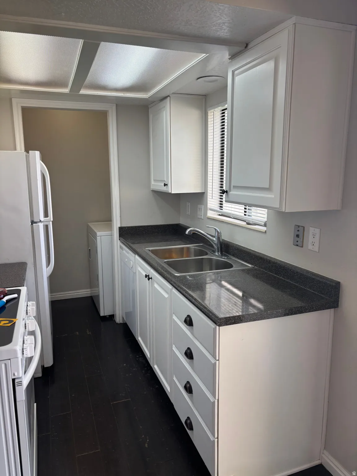White cabinetry with black hardware, dark wood-finish flooring, dark countertops, dual basin stainless steel sink, and a window with blinds