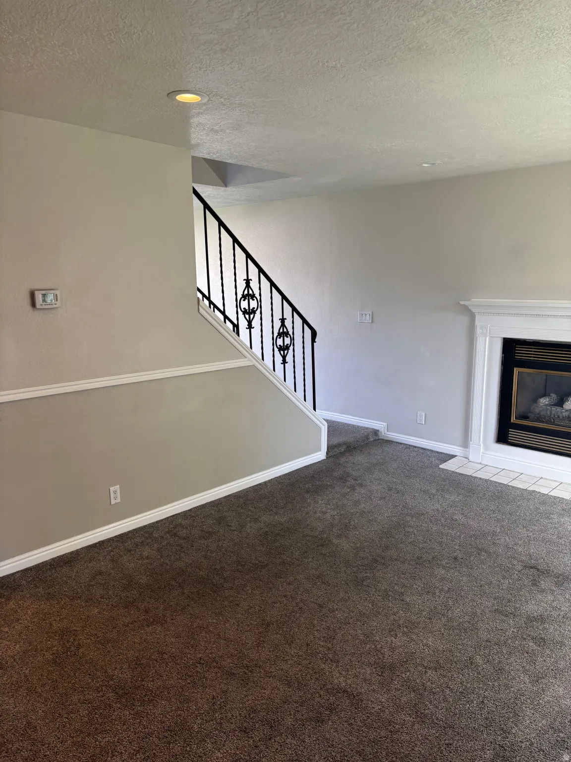 Recessed ceiling lighting, wrought iron staircase railing, carpeted flooring, and a fireplace with a white mantel and tile hearth