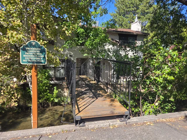 Wooden bridge with black wrought iron railings spanning a waterway, leading to a multi-level building with a visible chimney