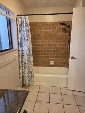 Bathtub with shower, brown square tile surround, and an adjacent window with blinds