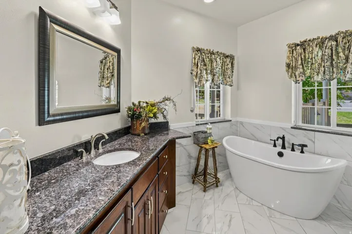 Freestanding soaking tub, dual windows, dark wood-finish vanity with an integrated sink, polished chrome faucet, and a large framed mirror