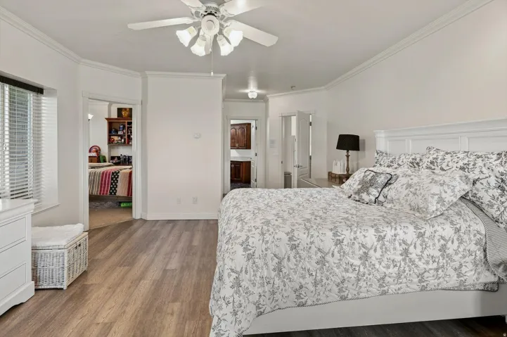 Wood-finish flooring, white paneled headboard, window with horizontal blinds, ceiling fan with integrated lighting, and crown molding
