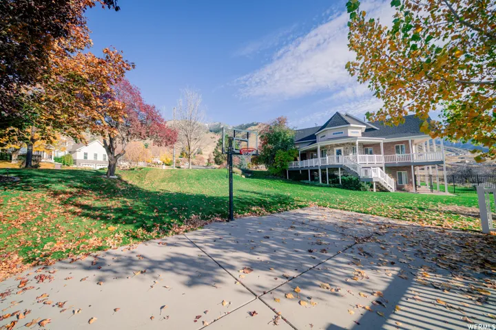 Expansive concrete pad, large grassy area, basketball hoop, and a two-story residence with a wraparound porch and white railings
