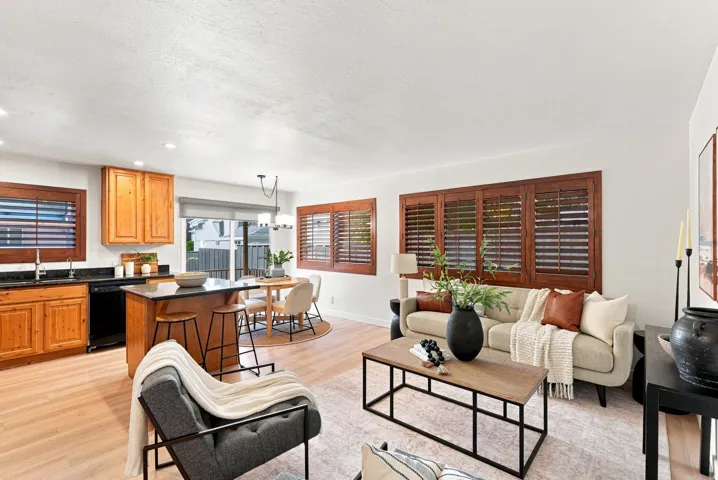 Open concept living area with wood-finish flooring, kitchen island with seating, dark countertops, and wooden plantation shutters
