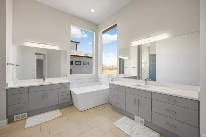 Dual vanities with gray cabinetry and white countertops, freestanding soaking tub, and tall windows
