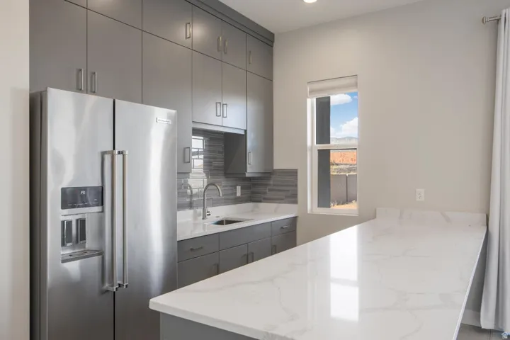 Stainless steel side-by-side refrigerator, gray flat-panel cabinetry, white stone-finish countertops, gray tile backsplash, and a stainless steel kitchen faucet