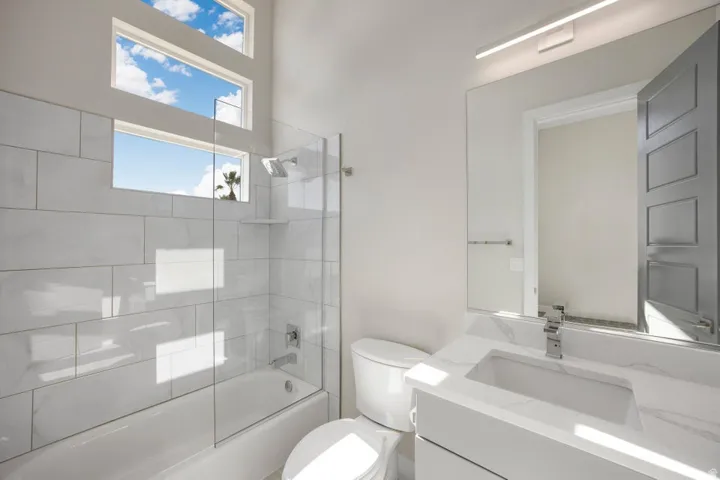 Bathroom featuring a white vanity with an integrated sink, polished chrome fixtures, a full-width mirror with linear overhead lighting, a white toilet, and a bathtub with a clear glass enclosure and subway tile surround