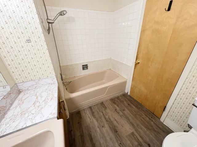 Shower-tub combination with white tile surround, wood-finish flooring, and a vanity featuring a marbled countertop