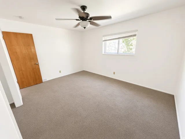 Neutral wall paint, textured carpeting, wooden interior door, window with blinds, ceiling fan with integrated lighting
