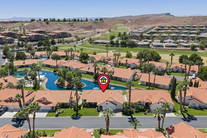 Residential architecture with red tile roofs, surrounded by mature palm trees and green landscaping, featuring a central water feature with small islands, and bordered by a golf course and distant arid hills