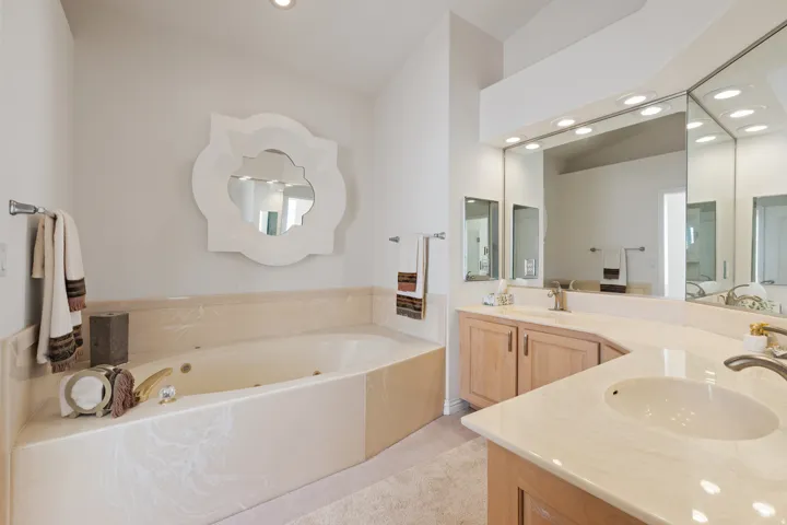 Recessed bathtub with integrated jets, dual vanity with undermount sinks, light-finish cabinetry, extensive vanity mirror, and overhead lighting