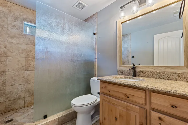 Textured glass shower partition, tile shower surround, wood-finish vanity with built-in storage, light-toned countertop, and framed mirror with integrated lighting