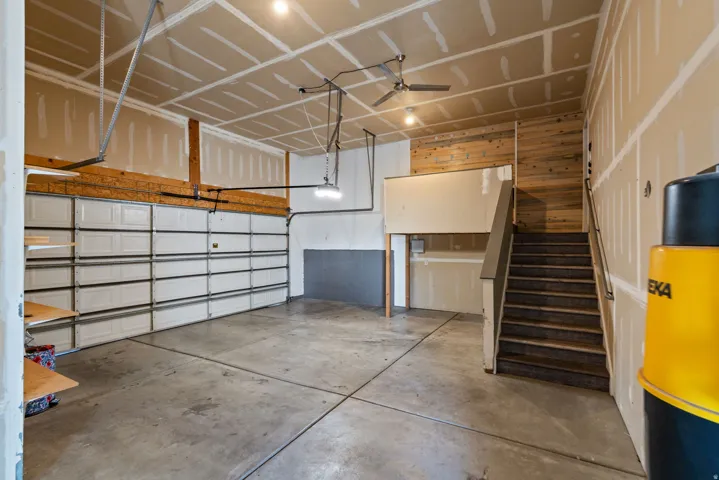 Two-car garage with concrete flooring, exposed wood beam detailing, ceiling-mounted light fixtures, and a staircase with a handrail