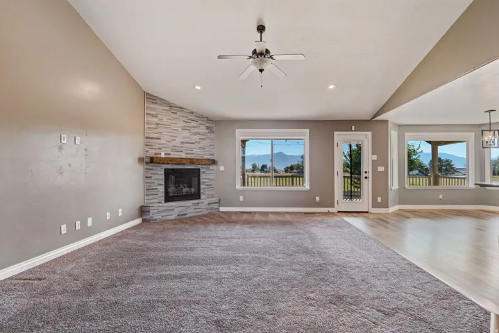 Vaulted ceiling, stone-surround fireplace with wood mantel, ceiling fan, recessed lighting, and expansive windows showcasing mountain scenery
