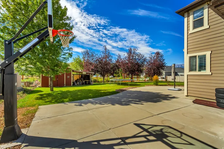 Expansive concrete patio with a basketball hoop, offering a broad grass lawn, mature trees, and a storage shed