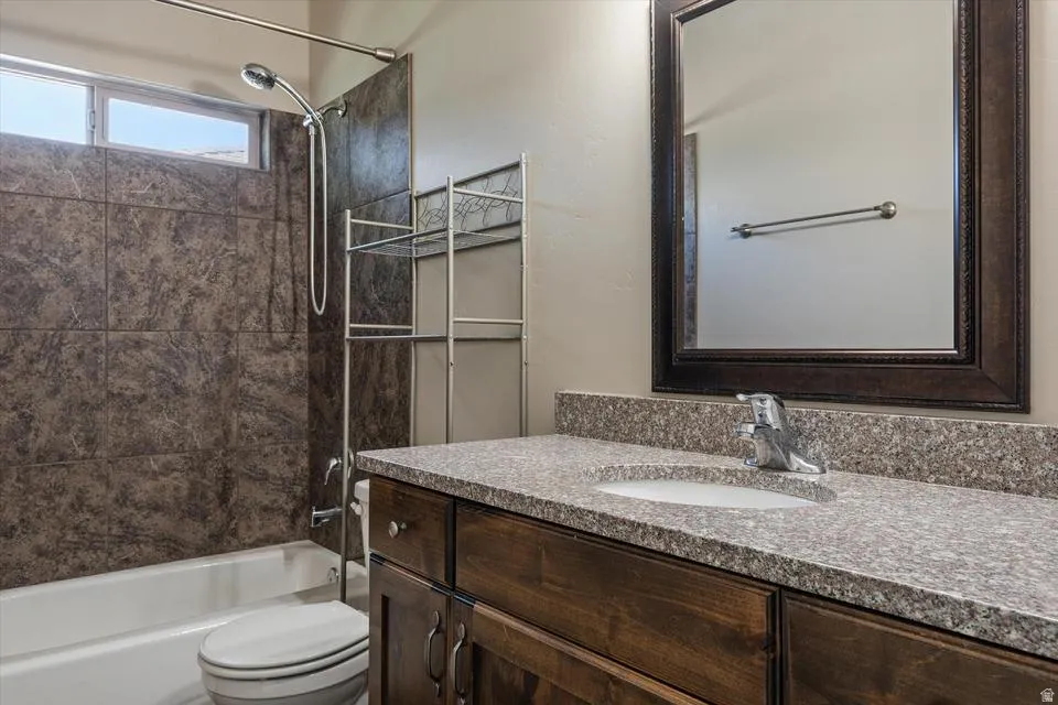 Brown tiled shower surround, white bathtub, wood-finish vanity, light-colored speckled countertop, and framed mirror