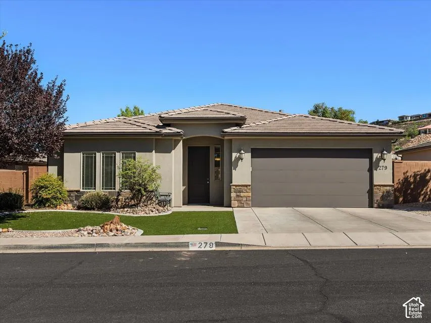 Stucco exterior with stone accents, tile roof, attached garage, concrete driveway, and low-maintenance artificial turf landscaping