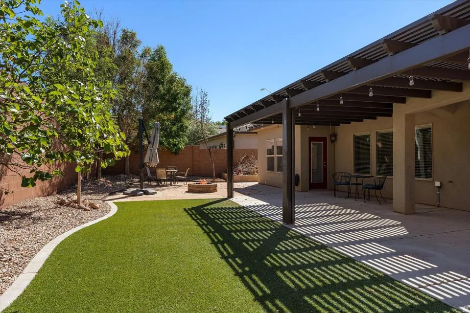 Expansive backyard featuring artificial turf, a built-in fire pit, a covered patio with a slatted pergola, and mature landscaping