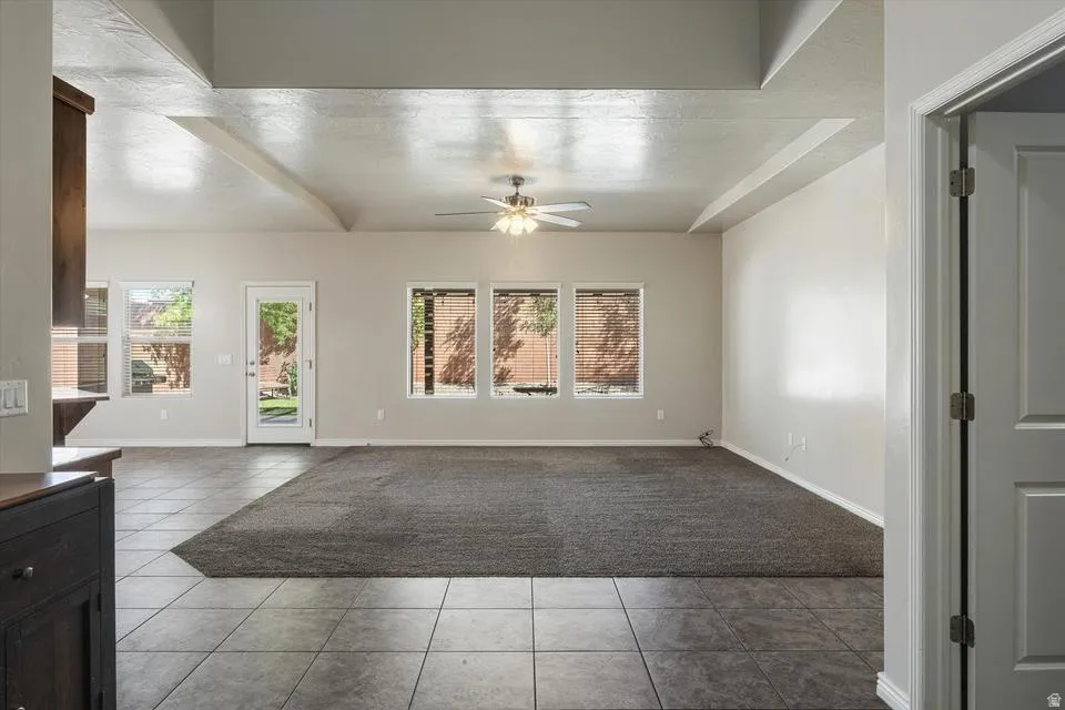 Tray ceiling with exposed beams, ceiling fan, multiple windows with blinds, tiled flooring, and carpeted area
