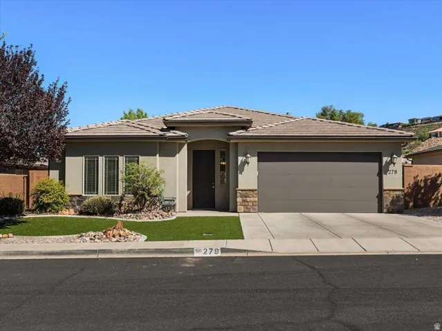 Stucco exterior with stone accents, tile roof, attached garage, concrete driveway, and low-maintenance artificial turf landscaping