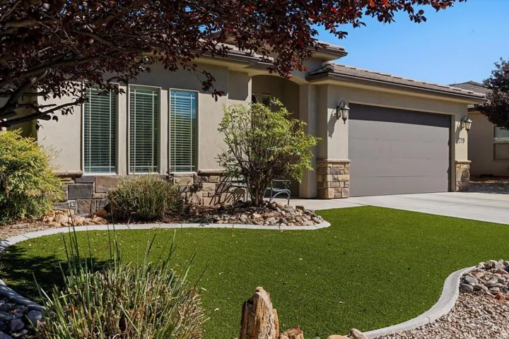 Stucco exterior with stone accents, covered entry, attached garage with decorative sconces, and artificial turf landscaping with rock beds