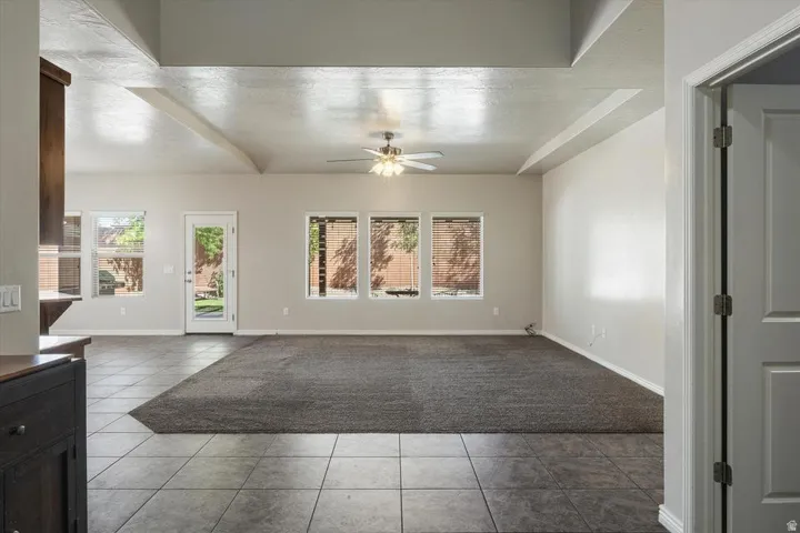 Tray ceiling with exposed beams, ceiling fan, multiple windows with blinds, tiled flooring, and carpeted area