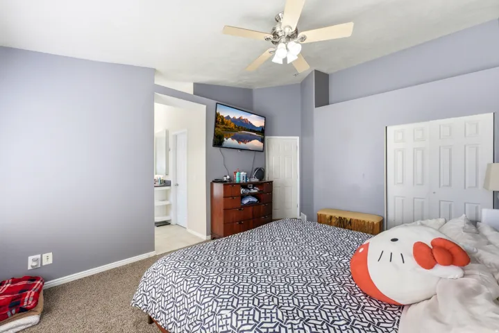 Neutral-toned carpeting and walls, ceiling fan with light fixture, bi-fold closet doors, and an en-suite bathroom with vanity