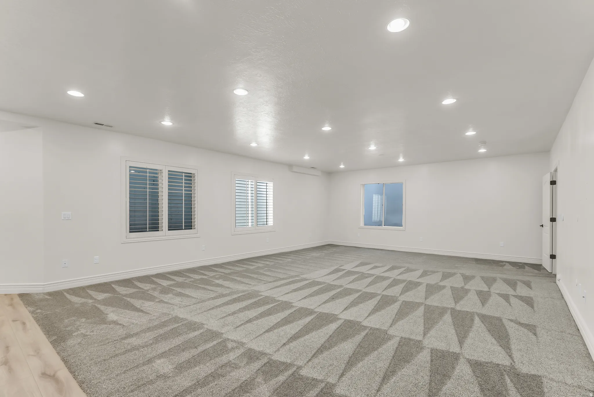Spacious room with recessed lighting, light wood-finish flooring, patterned carpet, and multiple windows with plantation shutters
