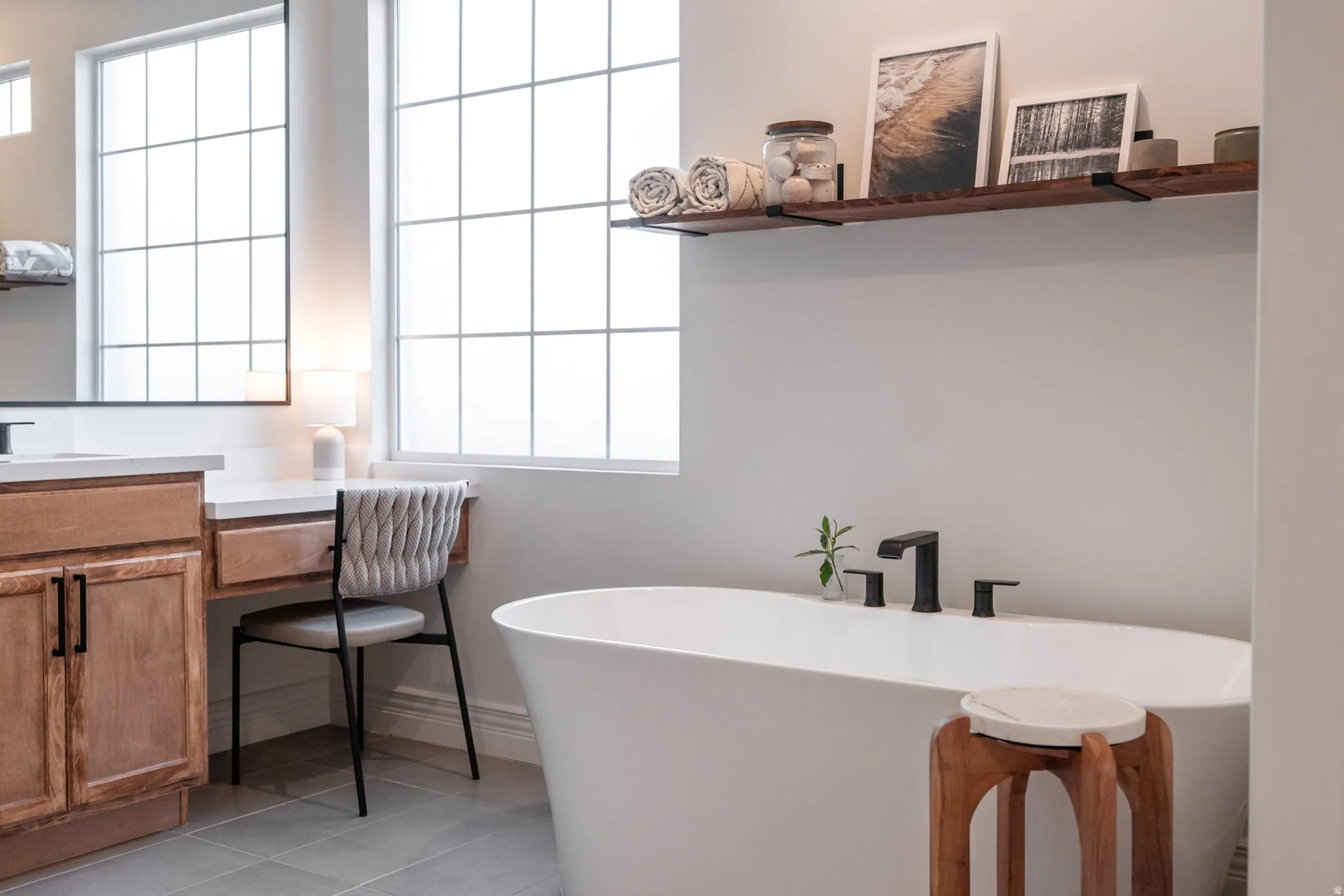 Freestanding soaking tub with matte black fixtures, grid-pattern windows, wood-finish floating shelf, and a built-in vanity desk