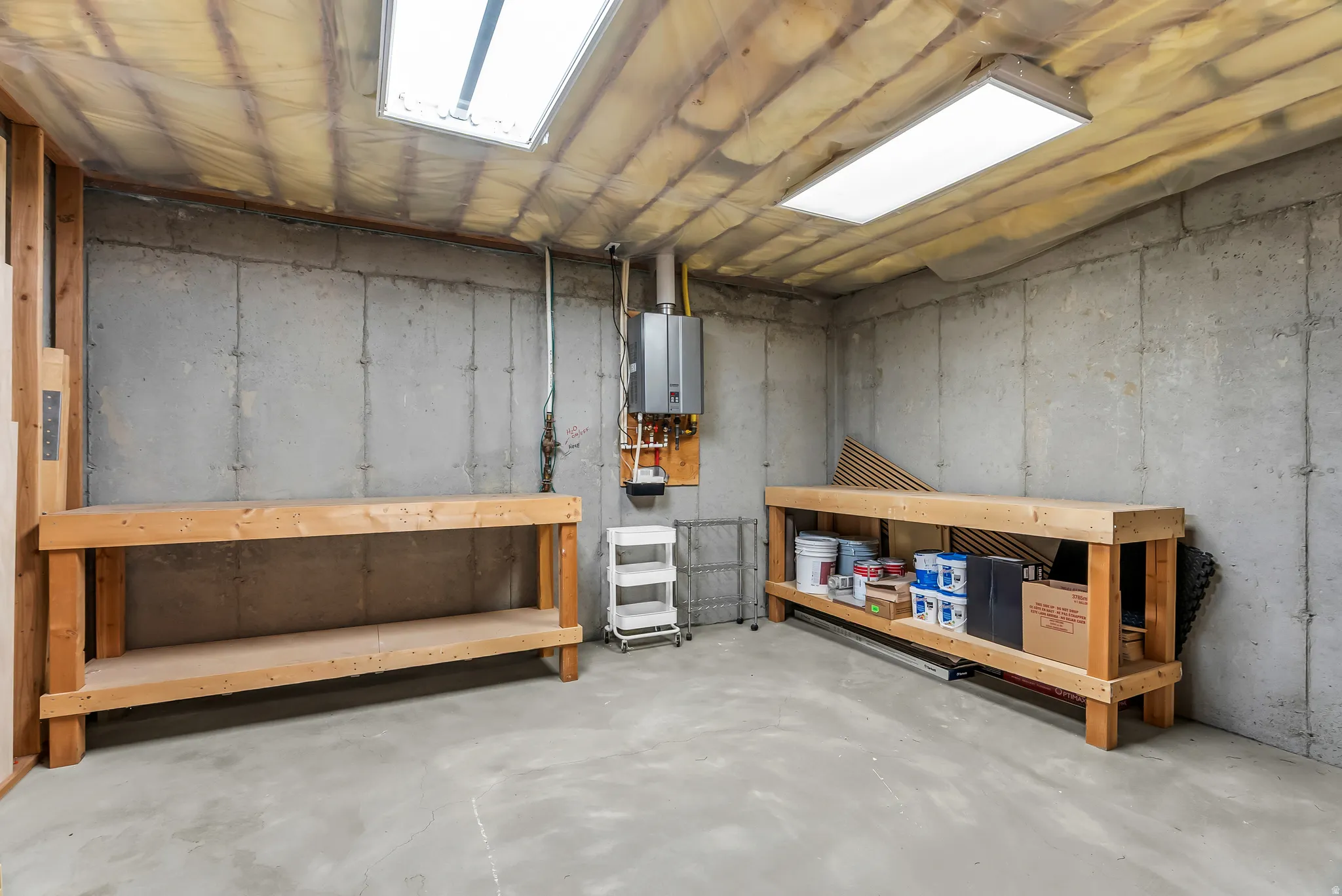 Concrete floor and walls, exposed ceiling insulation, dual overhead fluorescent lighting, and two built-in wooden workbenches